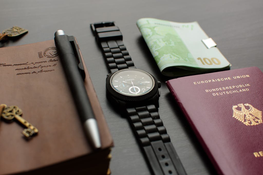 A neatly arranged flat lay depicting essential travel items such as a German passport, garmin watch, and euro bills on a desk.